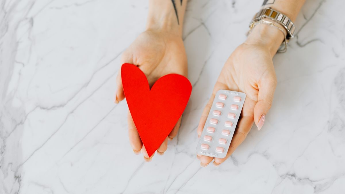 Close-up of hands holding a red heart shape and a blister pack of pills on a marble surface.